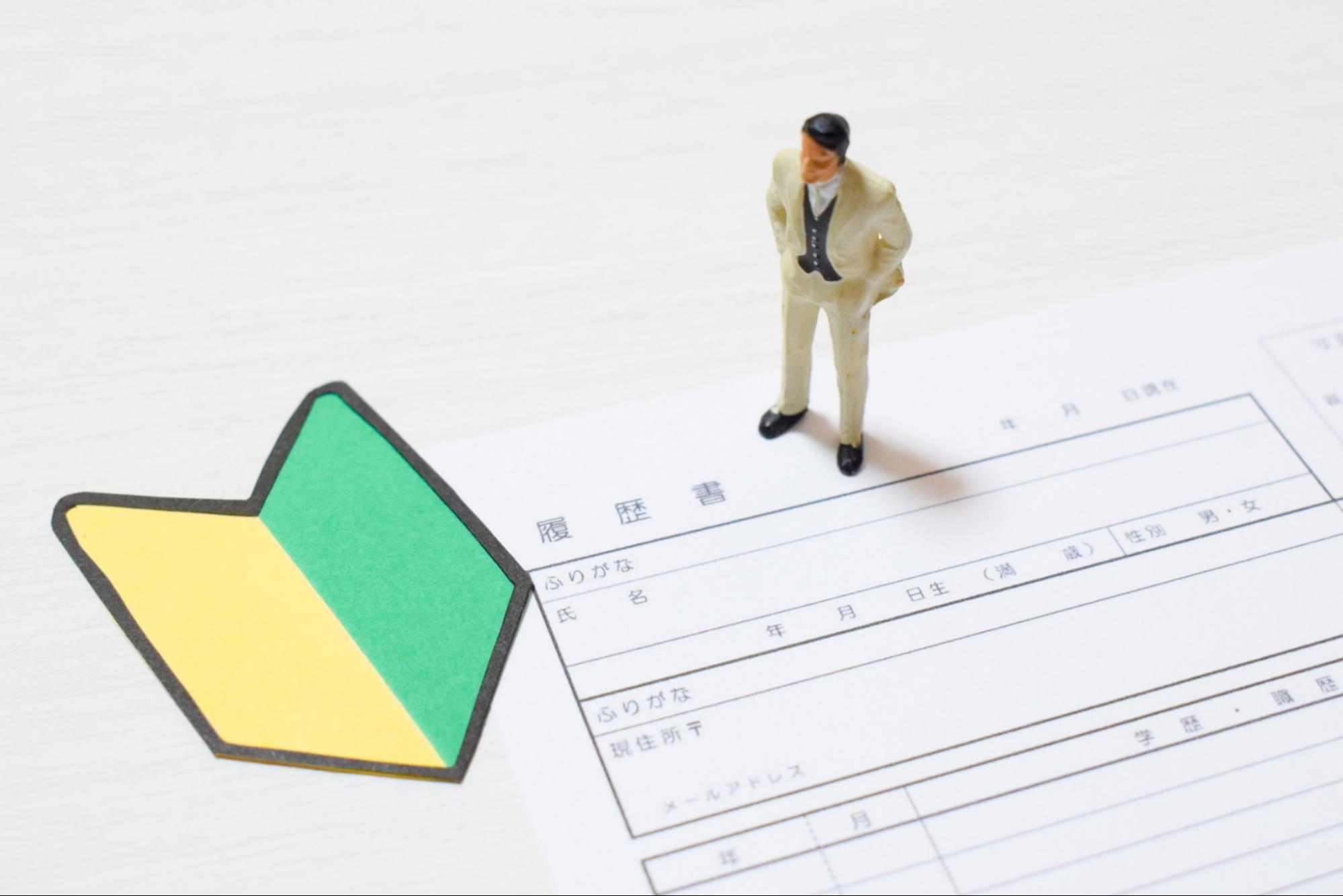 「履歴書」と書かれた用紙の上に初心者マークのプレートと男性のミニチュア人形が置かれている様子が写っている。