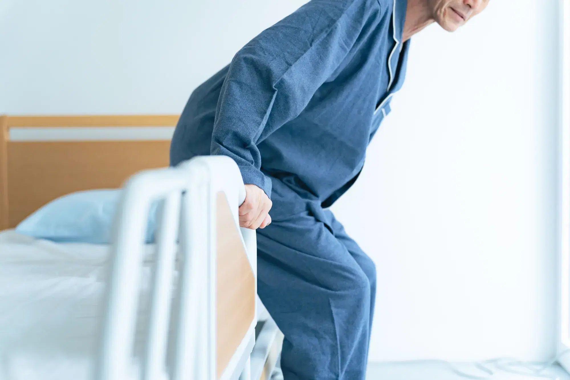 青いパジャマを着た男性がベッドの手すりをつかんで起き上がっている。