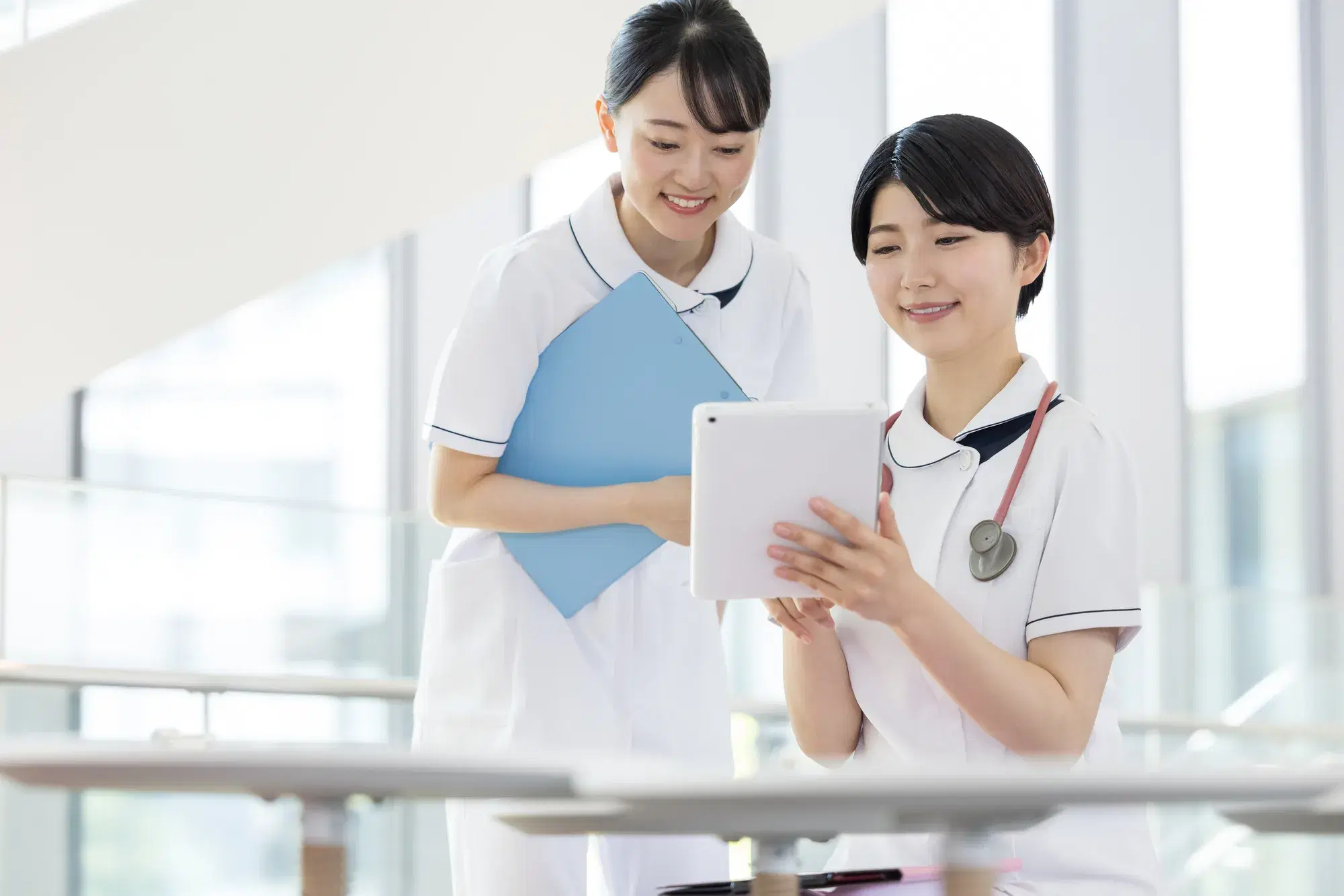 白衣を着た女性二人が白いタブレット端末をみながら話し合っている