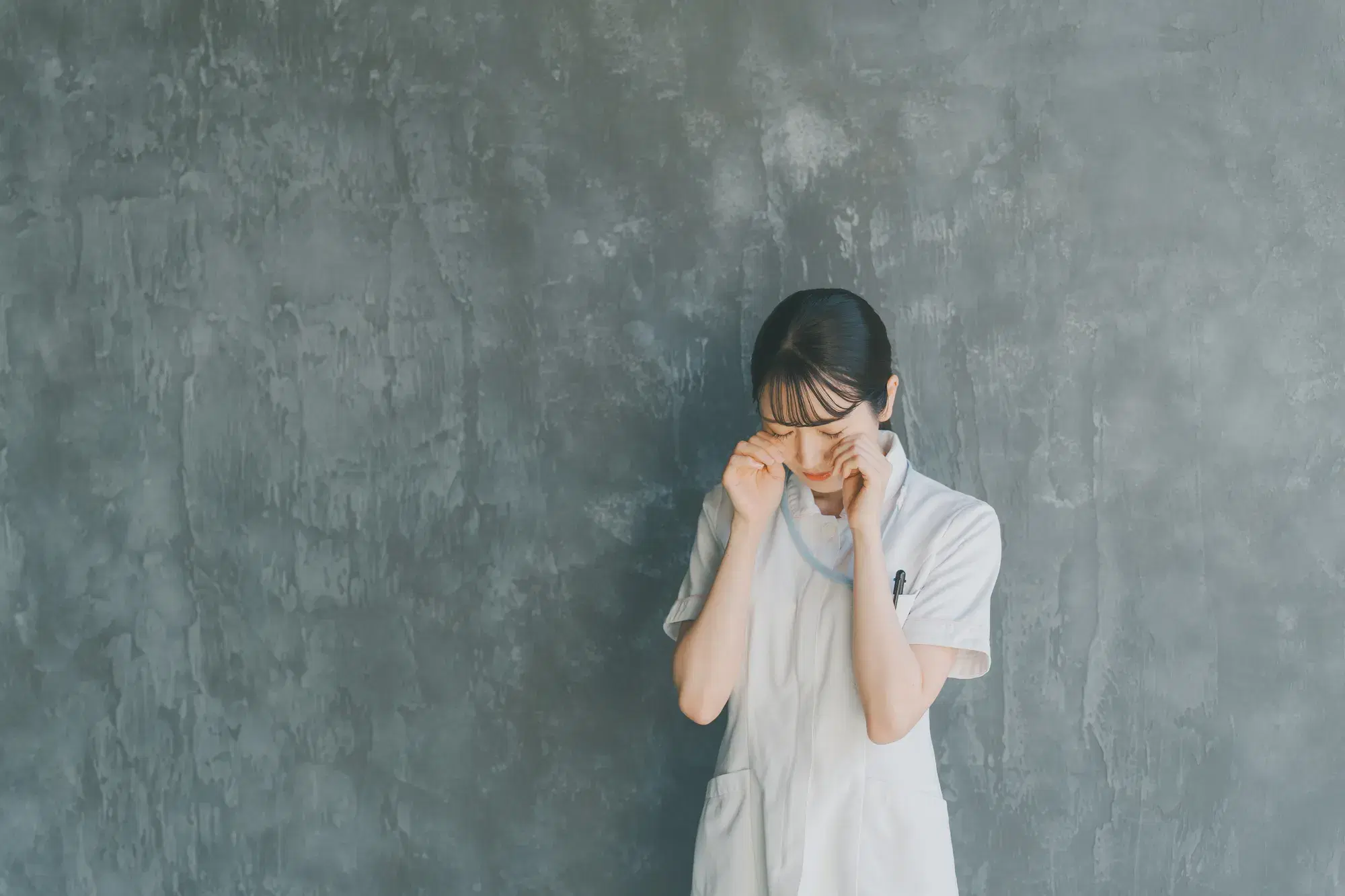 白い服を着た女性の医療従事者が両手で目をこすっている。