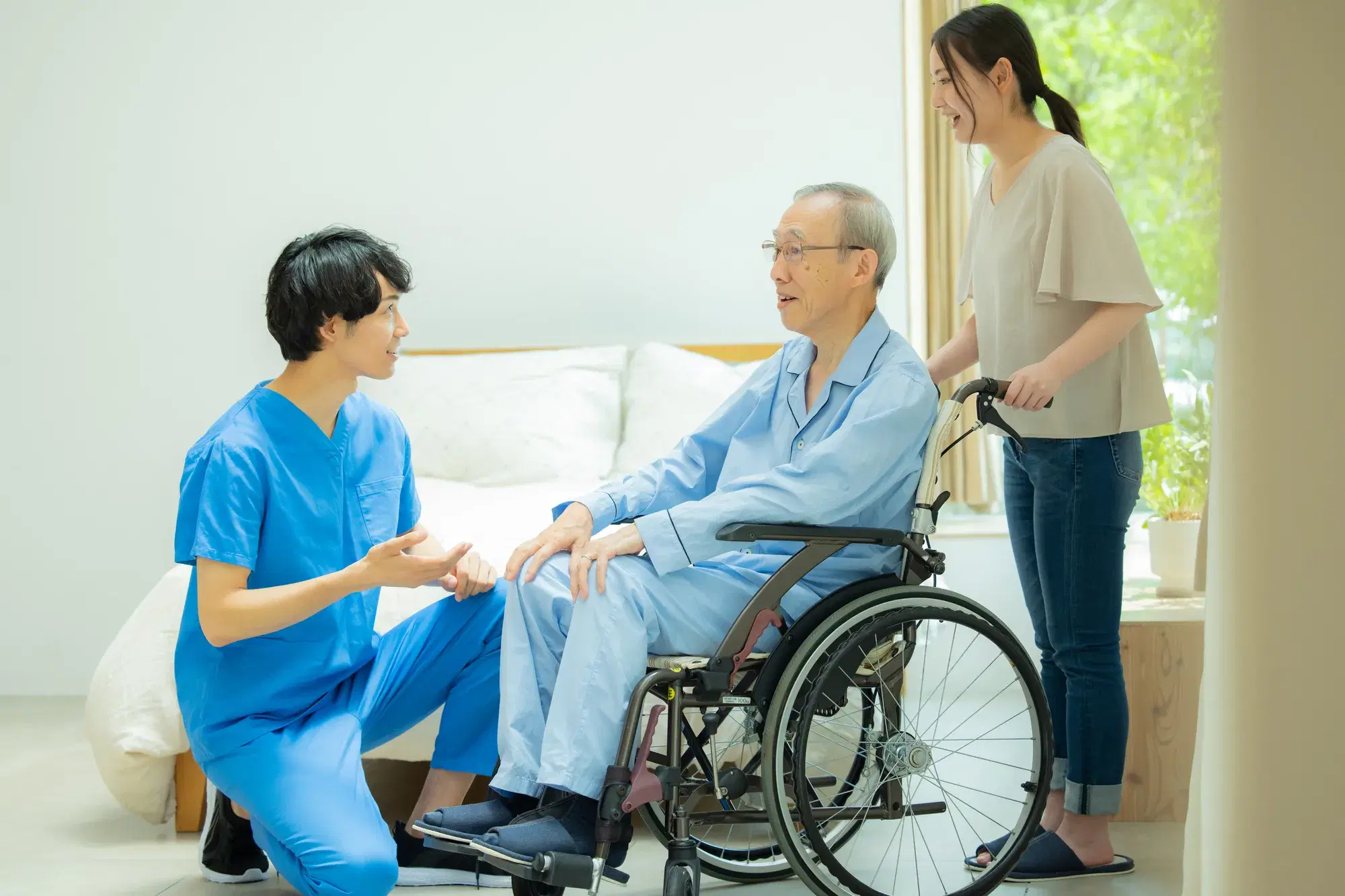青い服を着た老人がベージュの服を着た女性に乗っている車椅子を押されており男性の目の前には青い服を期着た医療関係の男性がいる