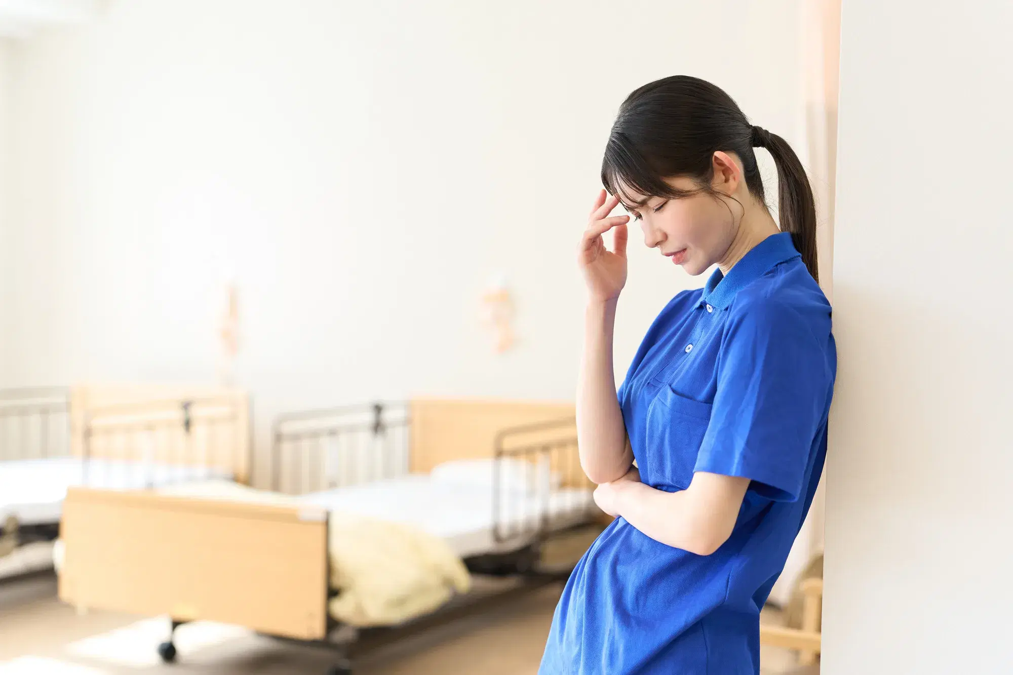 青い服を着た女性が介護施設のベッドがある部屋で壁にもたれかかっている