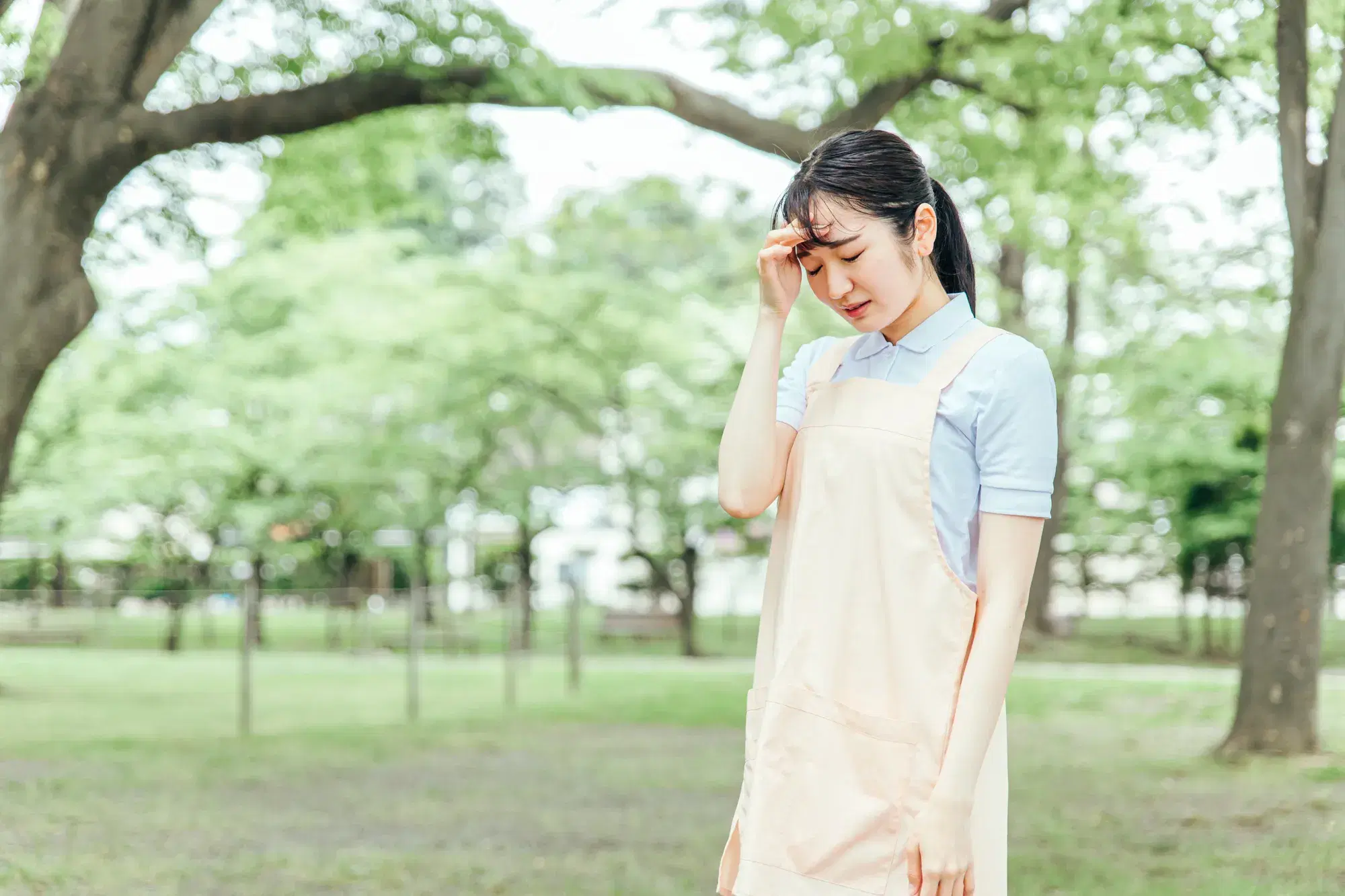 公園で頭に手を添えるピンク色のエプロンを着用した女性