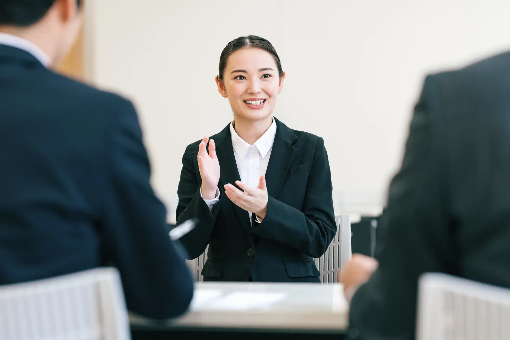 手前に黒いスーツを着た人物たちがおり、奥で黒いスーツを着た女性が手を叩いている。