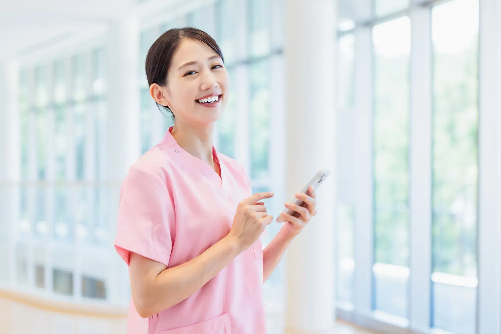 ピンクの服を着た医療従事者の女性がスマホを持って笑っている