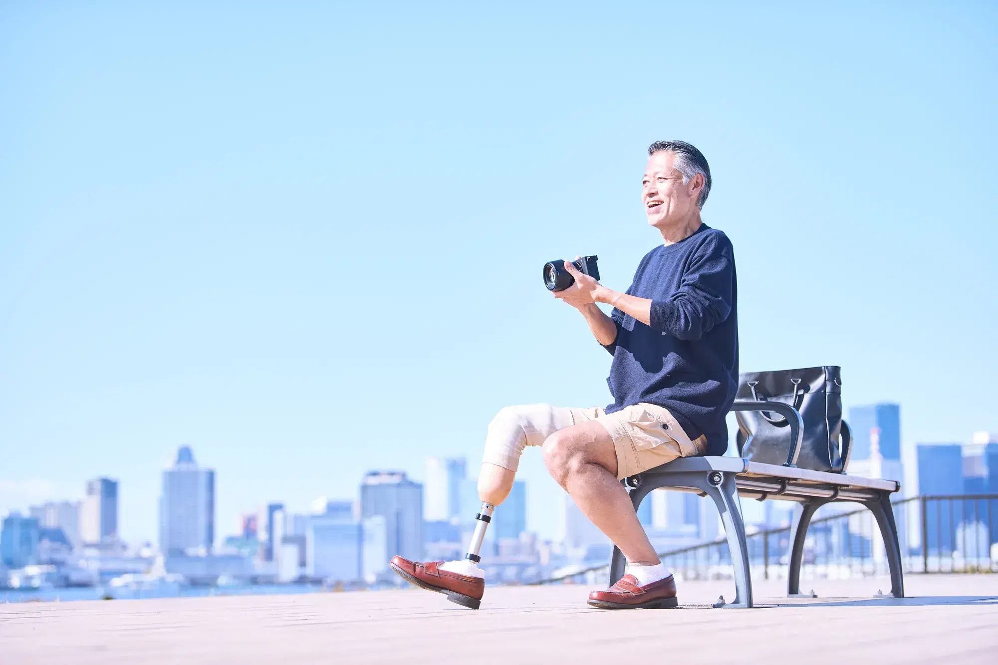 義足を付けた青い服を着た男性が椅子に座ってカメラを構えている