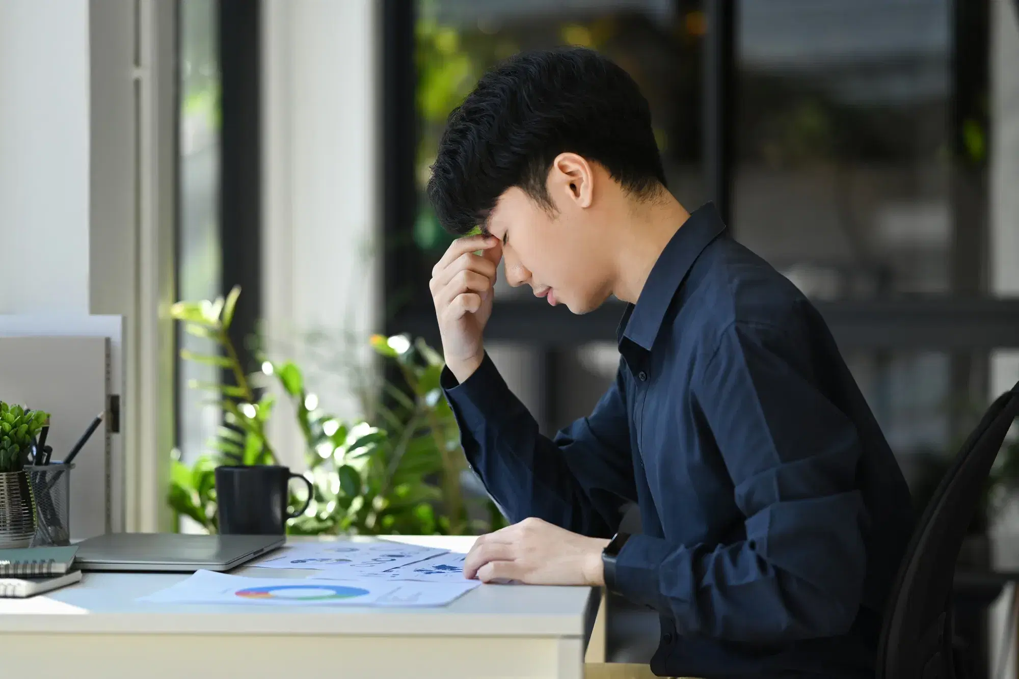 黒い服を着た男性が机の上に参考書を広げながら眉間をつまんでいる。