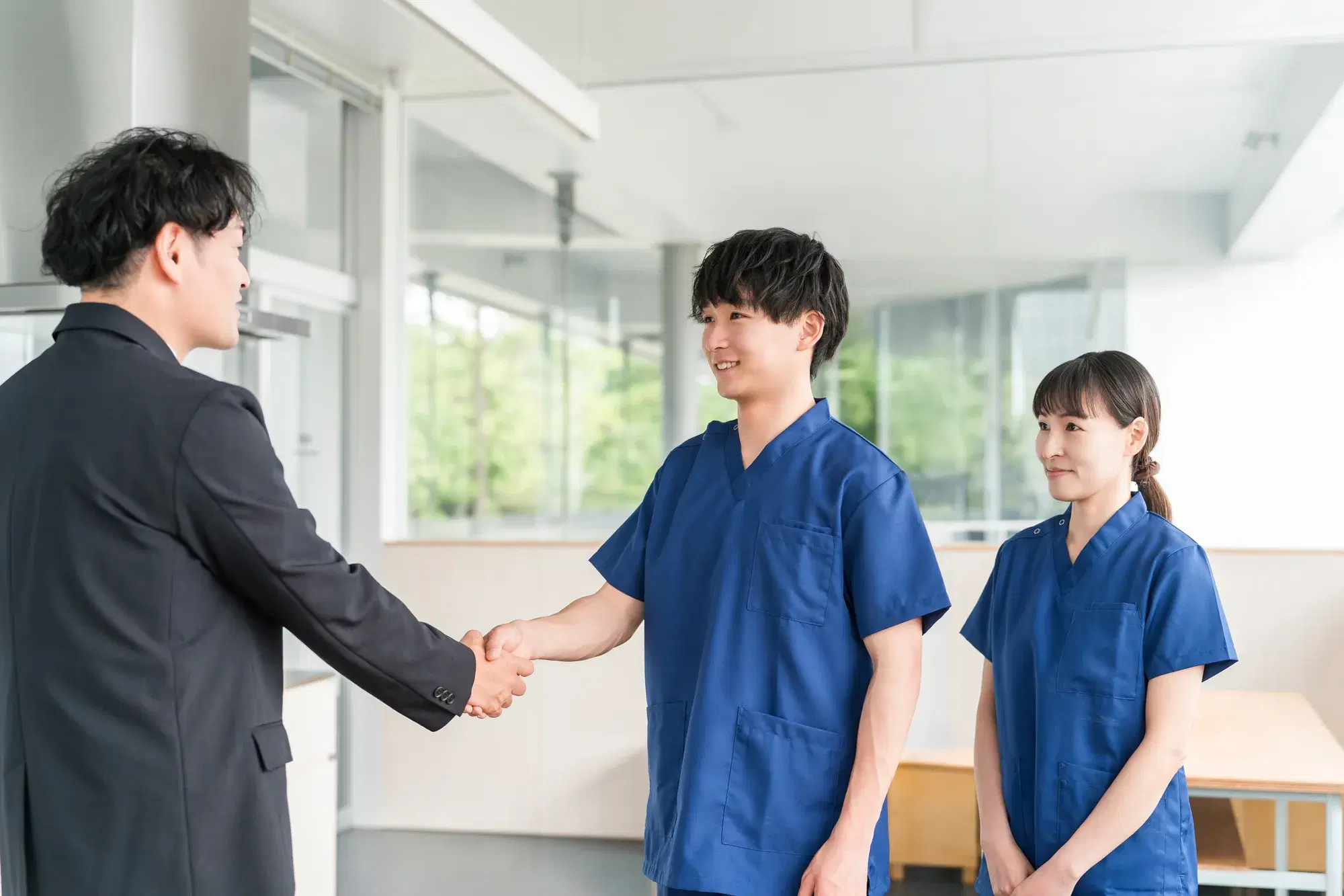 紺色のスクラブを着た男性と女性が右側におり、男性は黒いスーツを着た男性と握手をしている。