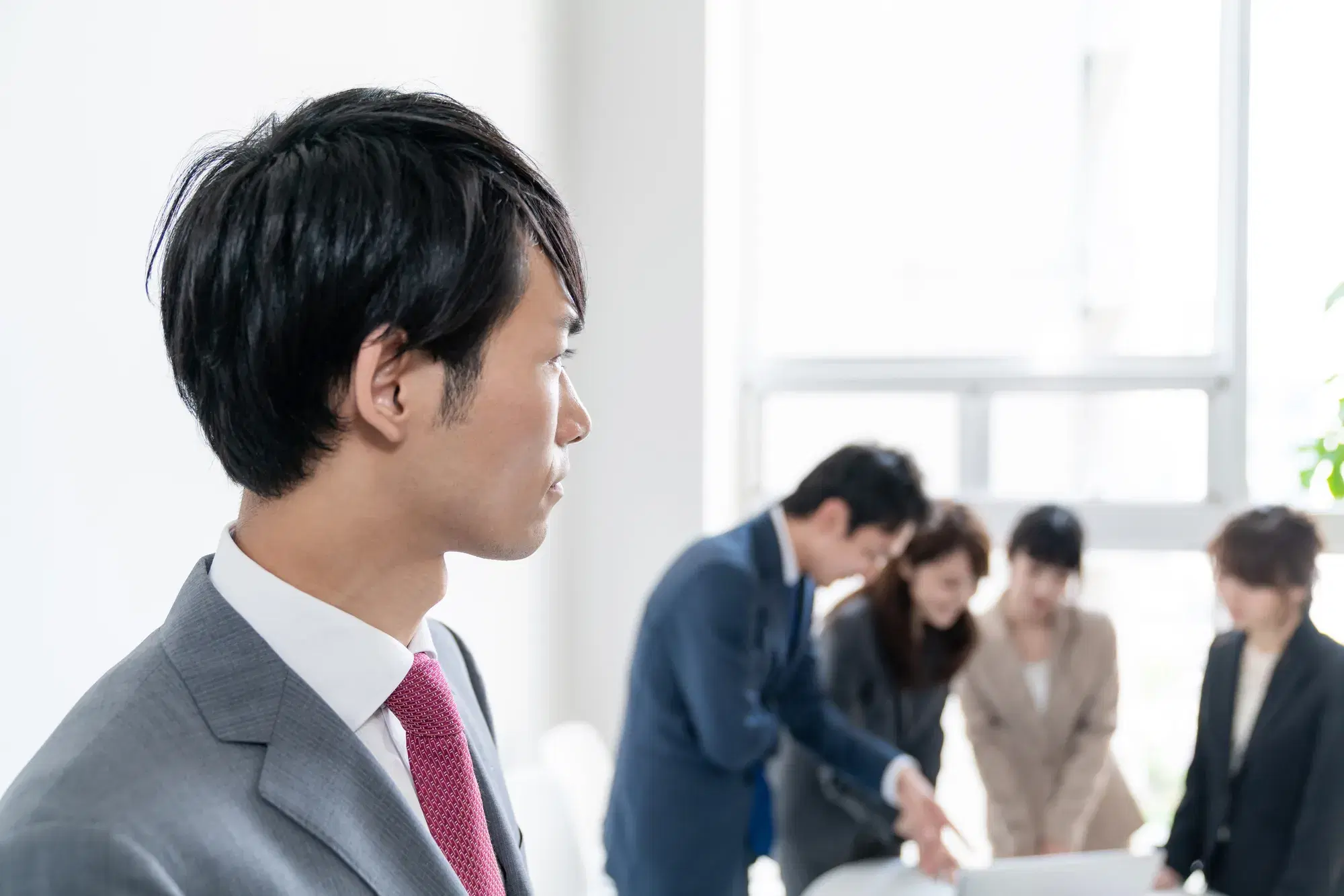 灰色のスーツを着た男性が同じ室内の固まっているグループを遠くから見る