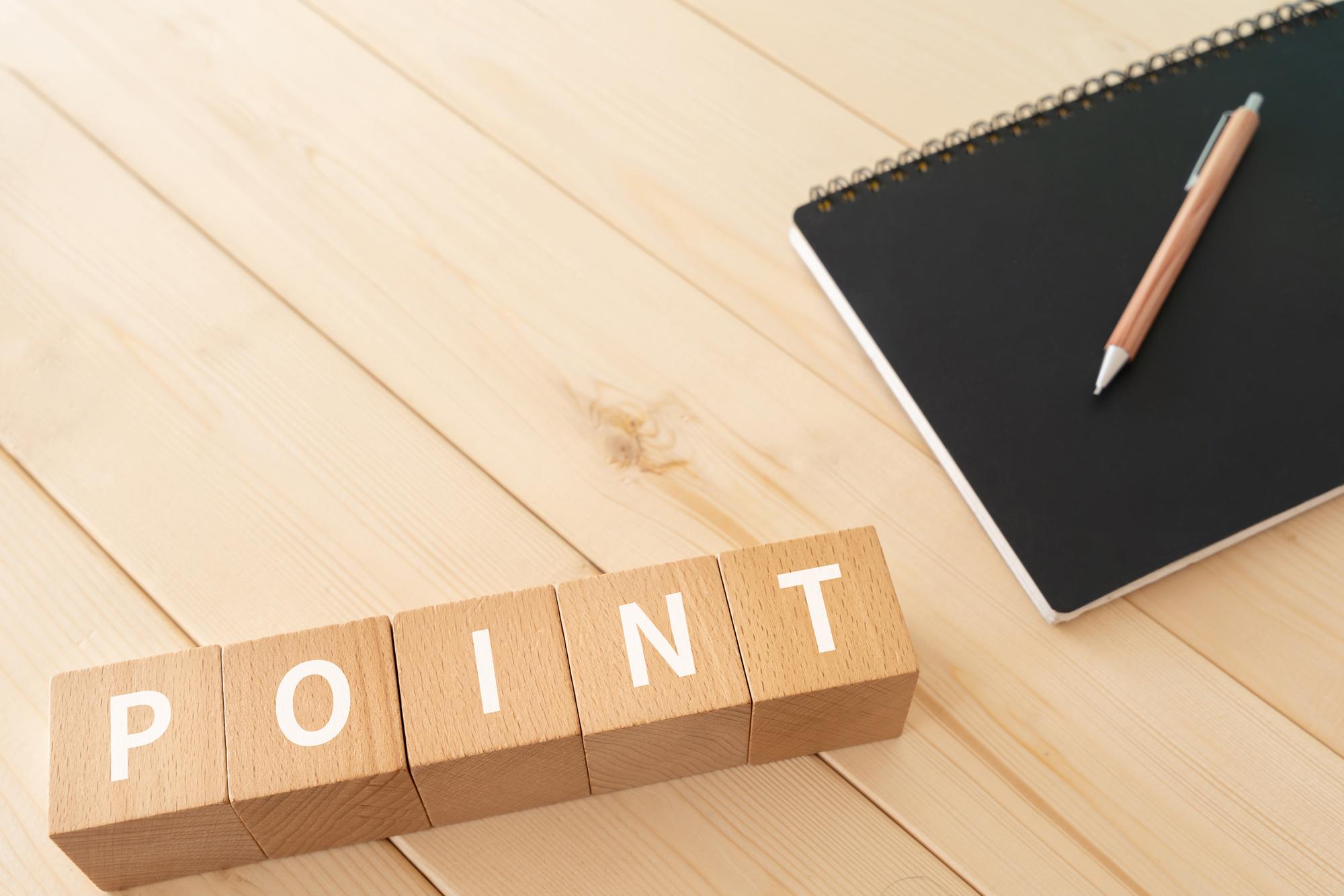 木目調のテーブルの上に「POINT」と書かれた木製ブロック、黒いノート、ペンが置かれている様子が写っている。